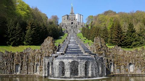 In einem Park eingebettet eine Wasserkaskade von unten fotografiert. Oben, am Beginn der Treppen, auf welchen Wasser runterfließt, steht eine Skulptur, ein überdimensionaler männlicher Körper.