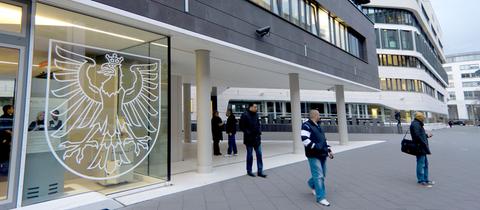 Menschen stehen vor einem Bürogebäude an dessen Fensterscheibe im Erdgeschoss das Frankfurter Wappen zu sehen ist.