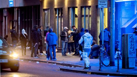 Menschen stehen am Abend im Frankfurter Bahnhofsviertels vor einem Drogenkonsumraum. Sie sind teilweise blau beleuchtet.