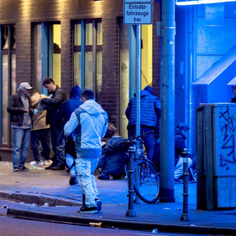 Menschen stehen am Abend im Frankfurter Bahnhofsviertels vor einem Drogenkonsumraum. Sie sind teilweise blau beleuchtet.