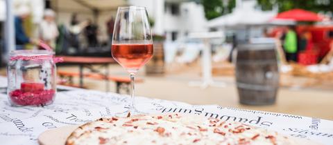 Ein Flammkuchen und ein stehen auf einem Tisch. Im Hintergrund zeigt sich eine Volksfest-Atmosphäre