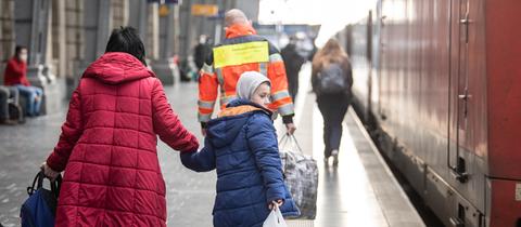Eine Frau geht mit ihrem Jungen an der Hand über den Bahnsteig, an dem ein Zug steht (von hinten fotografiert). Davor geht ein Mann einer Hilfsorganisation und trägt eine große Tasche.