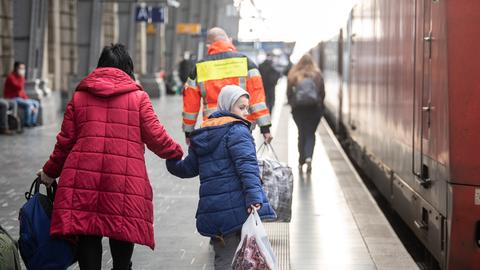 Eine Frau geht mit ihrem Jungen an der Hand über den Bahnsteig, an dem ein Zug steht (von hinten fotografiert). Davor geht ein Mann einer Hilfsorganisation und trägt eine große Tasche.