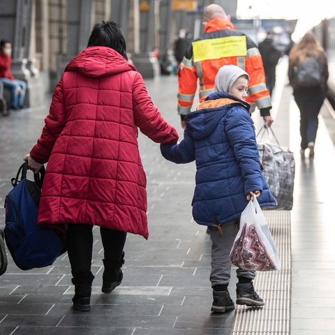 Eine Frau geht mit ihrem Jungen an der Hand über den Bahnsteig, an dem ein Zug steht (von hinten fotografiert). Davor geht ein Mann einer Hilfsorganisation und trägt eine große Tasche.