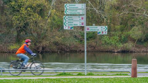 Ein Fahrradfahrer ist unterwegs an einem Fluss. Neben ihm sind Hinweisschilder auf Radwege zu sehen.