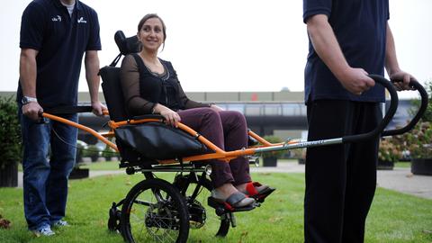 Der geländegängige Rollstuhl "Joëlette": Eine Frau sitzt in einem Rollstuhl, der wie eine Sänfte vorne und hinten von je einem Mann gehalten wird.