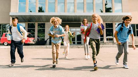 Sechs Kinder mit Schulranzen stuermen aus einem Schulgebäude, das hinter ihnen liegt.