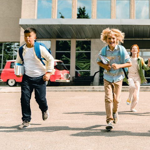 Sechs Kinder mit Schulranzen stuermen aus einem Schulgebäude, das hinter ihnen liegt.