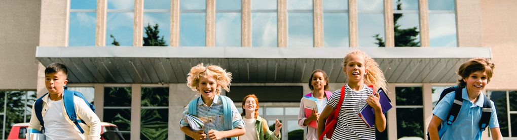 Sechs Kinder mit Schulranzen stuermen aus einem Schulgebäude, das hinter ihnen liegt.