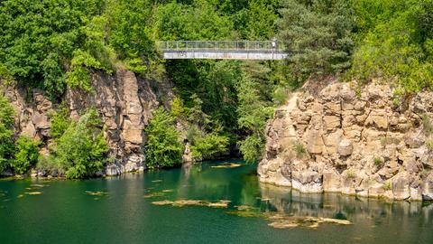 Eine Brücke zwischen zwei Felsen, darum stehen Bäume. Unten ist ein See. 