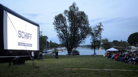 Das Foto zeigt eine Wiese. Links ist eine große Leinwand mit der Aufschrift "Schiff Film Festival" zu sehen, am rechten Bildrand Zuschauer in Plastikstühlen, die auf die Leinwand blicken. Im Hintergrund sieht man Wasser. 