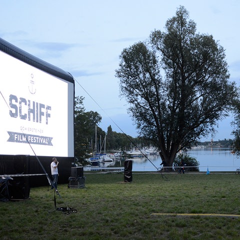 Das Foto zeigt eine Wiese. Links ist eine große Leinwand mit der Aufschrift "Schiff Film Festival" zu sehen, am rechten Bildrand Zuschauer in Plastikstühlen, die auf die Leinwand blicken. Im Hintergrund sieht man Wasser. 