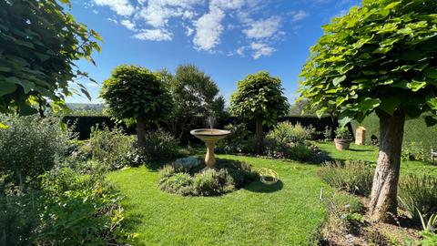 Ein Garten - darin steht ein Brunnen, drumherum sind diverse Bäume und Sträucher.