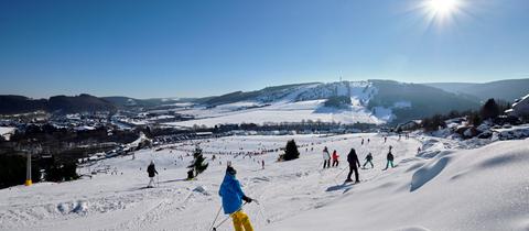 Mit Schnee bedeckte Landschaft bei strahlendem Sonnenschein. Menschen fahren Ski.