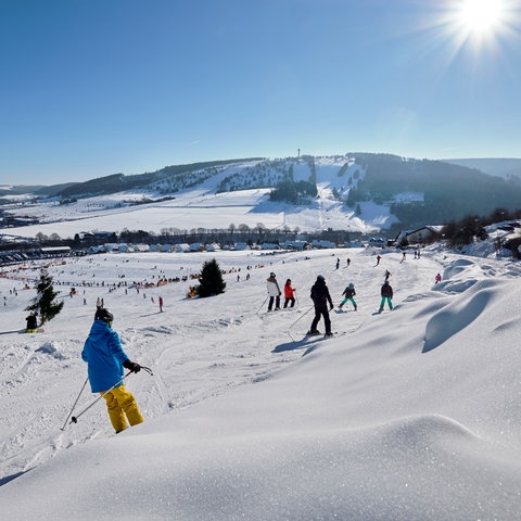 Mit Schnee bedeckte Landschaft bei strahlendem Sonnenschein. Menschen fahren Ski.