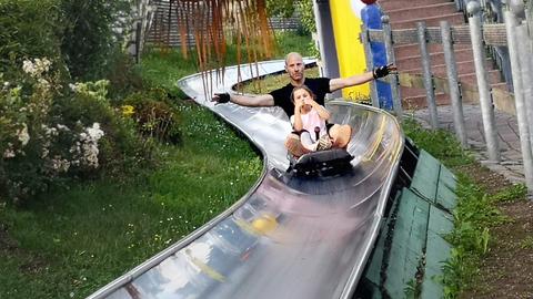 Ein Mann und ein Kind hocken in einer Art Schlitten und rutschen eine "Metallbahn" herunter. Der Mann breitet seine Arme aus. Drumherum viel Grün und farbenfrohe Bauten. 