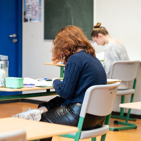 Drei Schülerinnen sitzen mit gebeugten Oberkörpern an Tischen in einem Klassenzimmer.