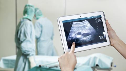 Foto: Im Vordergrund hält eine Hand ein Tablet mit einer Ultraschalldarstellung, im Hintergrund unscharf zwei Menschen in grüner medizinischer Schutzkleidung und eine Liege.