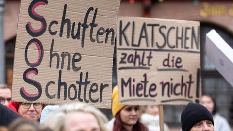 Demonstranten halten Schilder „Schuften ohne Schotter“ und „Klatschen zahlen die Miete nicht!“ hoch.