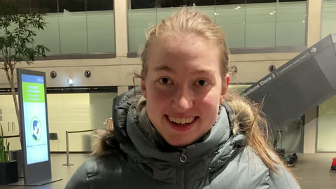 Eine junge Frau in Daunenjacke steht in einer Halle und lächelt in die Kamera.