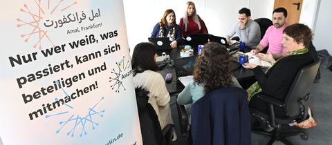 Das Bild zeigt einen schwarzen Holztisch, an dem sieben Frauen und Männer sitzen. Sie haben Laptops und Notizbücher vor sich. Links im Bild ist ein Banner zu sehen, auf dem steht: Nur wer weiß, was passiert, kann sich beteiligen und mitmachen!