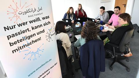 Das Bild zeigt einen schwarzen Holztisch, an dem sieben Frauen und Männer sitzen. Sie haben Laptops und Notizbücher vor sich. Links im Bild ist ein Banner zu sehen, auf dem steht: Nur wer weiß, was passiert, kann sich beteiligen und mitmachen!