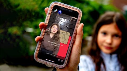 Hand mit einem Smartphone auf dessen Screen ein Video zu sehen ist (scharf im Vordergrund). Im Hintergrund (unscharf) ein junges Mädchen, welches das Smartphone hochhält.