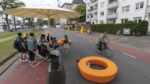 Der Mainkai ist autofrei, eine Gruppe von Menschen hat sich unter einem Sonnenstuhl versammelt, eine Person sitzt auf einem Liegestuhl. 