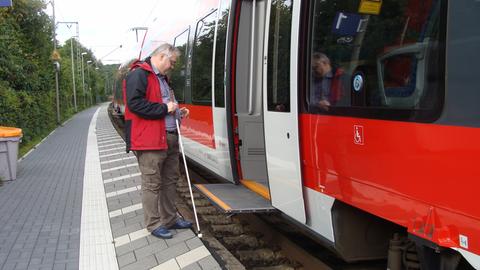 Ein sehbehinderter Mann mit Blindenstock steht vor einer offenen Zugtür. Vor ihm klafft eine Lücke zwischen Zug und Bahnsteigkante.