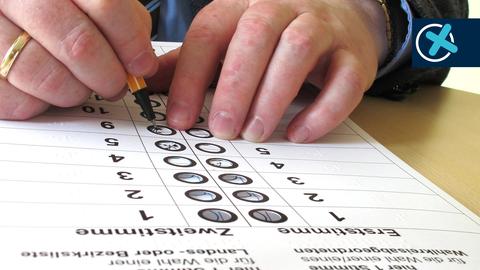 Nahaufnahme von Händen, die mit einem Stift einen Wahl-Stimmzettel ausfüllen. Zwischen Stift und Wahlzettel liegt eine Schablone mit Löchern und sehbehindertengerechter Beschriftung.