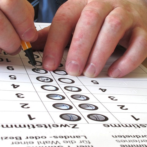 Nahaufnahme von Händen, die mit einem Stift einen Wahl-Stimmzettel ausfüllen. Zwischen Stift und Wahlzettel liegt eine Schablone mit Löchern und sehbehindertengerechter Beschriftung.