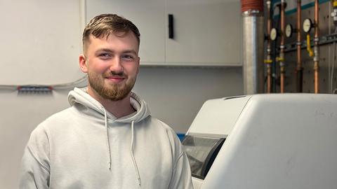 Portrait eines jungen Mannes, der in einem Werkraum steht und in die Kamera lächelt.