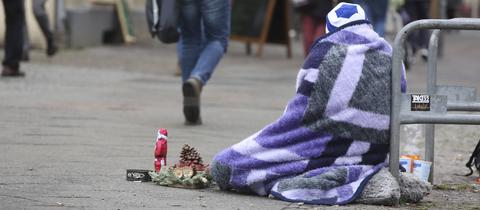 Wohnungsloser Mann sitzt mit einer kleinen Weihnachtsdeko, eingehüllt in eine Decke, auf dem Boden eines Einkaufszentrums.