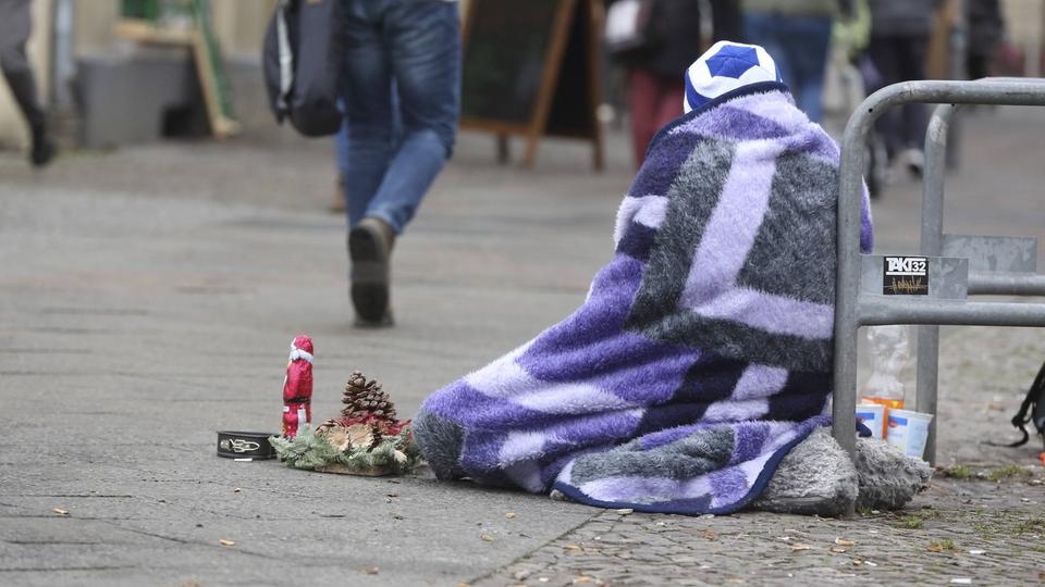 Wohnungsloser Mann sitzt mit einer kleinen Weihnachtsdeko, eingehüllt in eine Decke, auf dem Boden eines Einkaufszentrums.