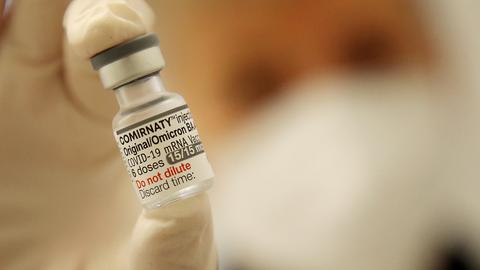 Im Bildvordergrund eine Ampulle mit dem Corona-Impfstoff von BioNTech/Pfizer in Großaufnahme, gehalten von Daumen und Zeigefinder (Unscharf). Im Hintergund unscharf ein maskiertes Gesicht.