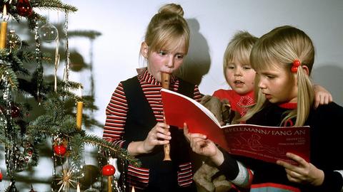 Zwei singende Mädchen und eins mit Blockflöte neben einem Weihnachtsbaum