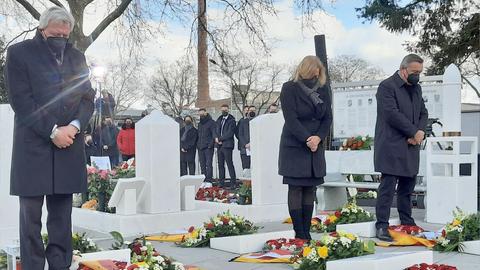 Ministerpräsident Bouffier, Bundesinnenministerin Faeser, OB Kaminsky und weitere Menschen stehen an Gräbern auf deinem Friedhof und gedenken mit gesenkten Köpfen.
