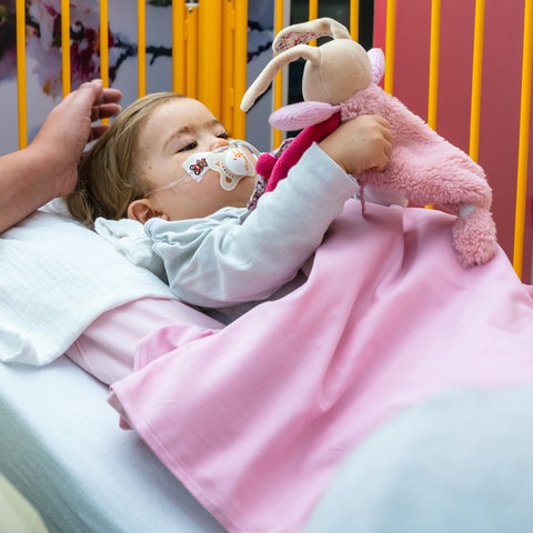 Ein Kleinkind liegt mit Schnuller, Atemschlauch und Stofftier im Bett. Zwei Frauen mit Masken beugen sich zu ihm.