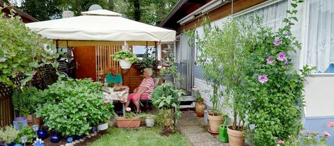 Ein älterer Mann und eine ältere Frau sitzen an einem Tisch unter einem Pavillonzeltdach auf einer Terrasse. Drumherum viele Pflanzen, ein Wohnwagen und ein Anbau.