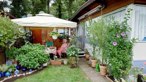 Ein älterer Mann und eine ältere Frau sitzen an einem Tisch unter einem Pavillonzeltdach auf einer Terrasse. Drumherum viele Pflanzen, ein Wohnwagen und ein Anbau.