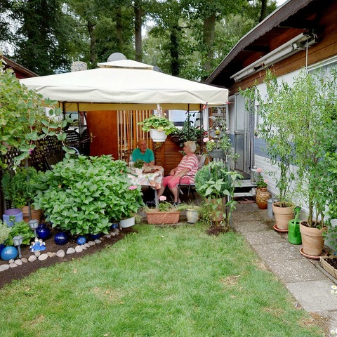 Einälterermann und Einältererfrau Sa Einem tisch unerem pavillonzeltdach aufgerer Terrasse. Drumherum Viele Pflanzen, Ein Wohnwagen und Ein Anbau.