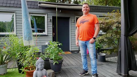 Ein Mann in orangefarbenem T-Shirt steht auf einer Terrasse mit vielen Pflanzen und Dekorationsobjekten. Im Hintergrund ein kleines Wohnhaus.