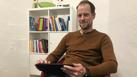 Der Erziehungswissenschaftler Timo Noll sitz mit einem Tablet auf einem orangenen Sessel. Im Hintergrund ist ein weißes Regal mit vielen Büchern zu sehen.