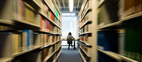 Eine Studentin sitzt mit ihrem Notebook in der Zentralbibliothek der Universität. Rechts und links sieht man hohe Regale, in denen unzählige Bücher stehen.