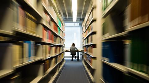Eine Studentin sitzt mit ihrem Notebook in der Zentralbibliothek der Universität. Rechts und links sieht man hohe Regale, in denen unzählige Bücher stehen.
