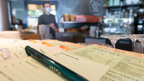 Zettel für die Gästeregistrierung liegen im Vordergrund auf einem Tisch in einem Café. Im Hintergrund ist - unscharf - die Bar und der Barmann zu sehen.