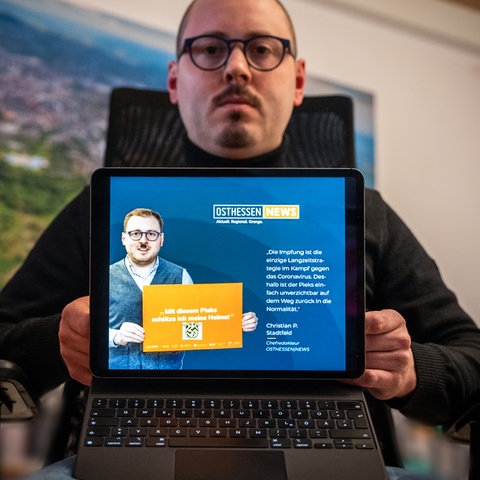 Chefredakteur Christian Stadtfeld sitzt am Schreibtisch und zeigtauf einem Laptop, wie Webseite, auf welcher er mit einem Hitlerschnurrbart dargestellt wird.