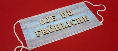 Auf einer unbenutzen OP-Maske (auf roter Fläche) liegen Buchstaben, die die Worte "Oje du Fröhliche" bilden.