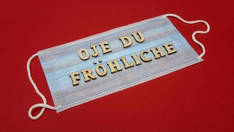Auf einer unbenutzen OP-Maske (auf roter Fläche) liegen Buchstaben, die die Worte "Oje du Fröhliche" bilden.