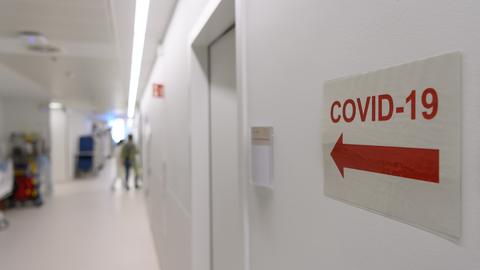 Ein Schild mit einem Richtungspfeil und der Aufschrift "Covid-19" Bereich klebt in einem Klinikflur an der Wand. 